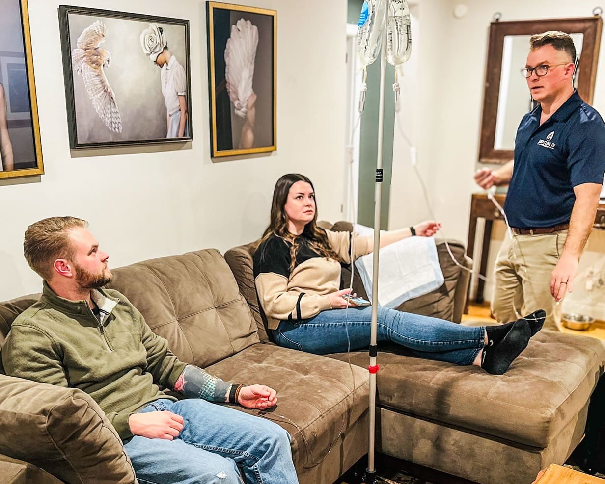 Three people in a casual living room setting, one appears to be giving a presentation or explaining something to the other two who are seated on a brown sofa, while a mobile IV hydration stand suggests a healthcare