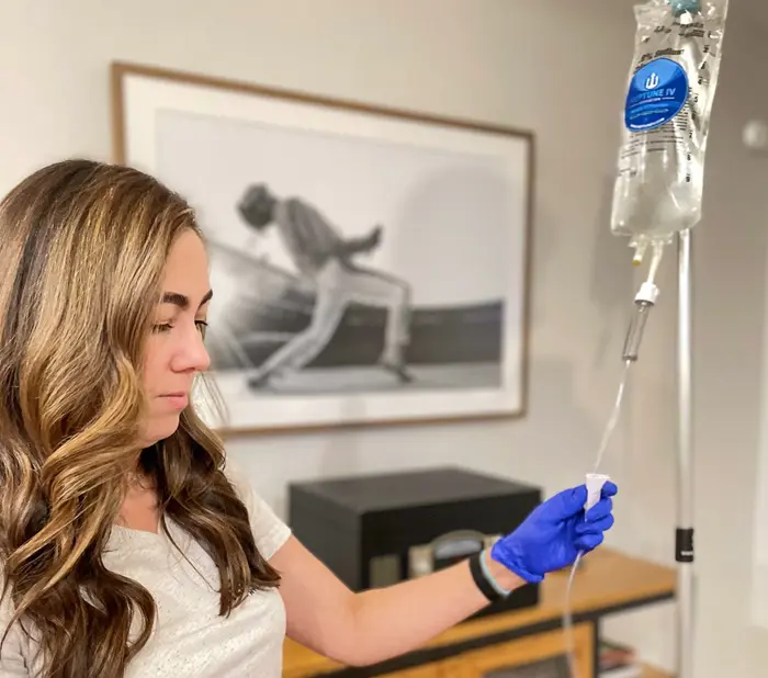 A woman in a white shirt and wearing blue gloves is adjusting an iv drip for mobile iv therapy in a home setting, with a black and white framed artwork in the background.